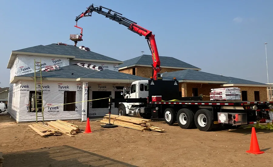A knuckle-boom crane truck lifting roofing materials to a house under construction
