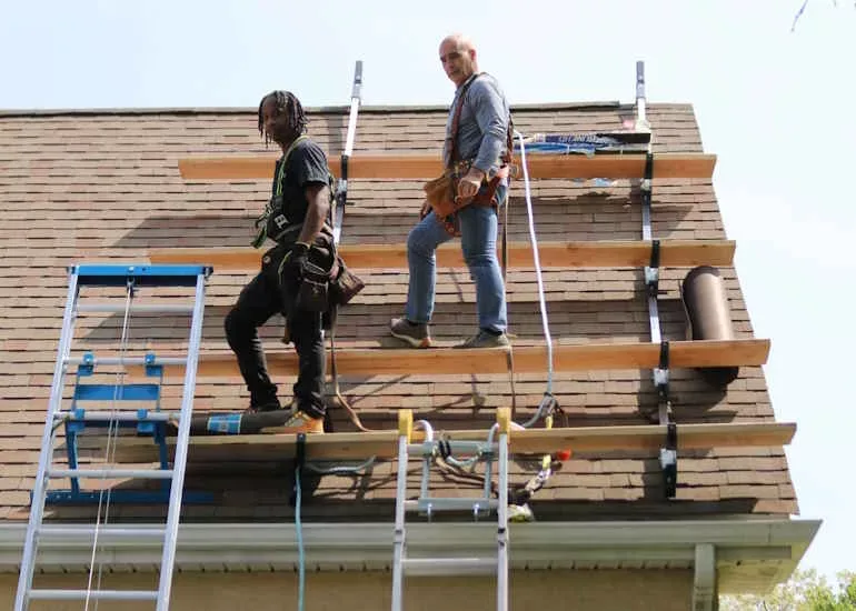 Two roofers on scaffolding tearing off and replacing shingles on a house