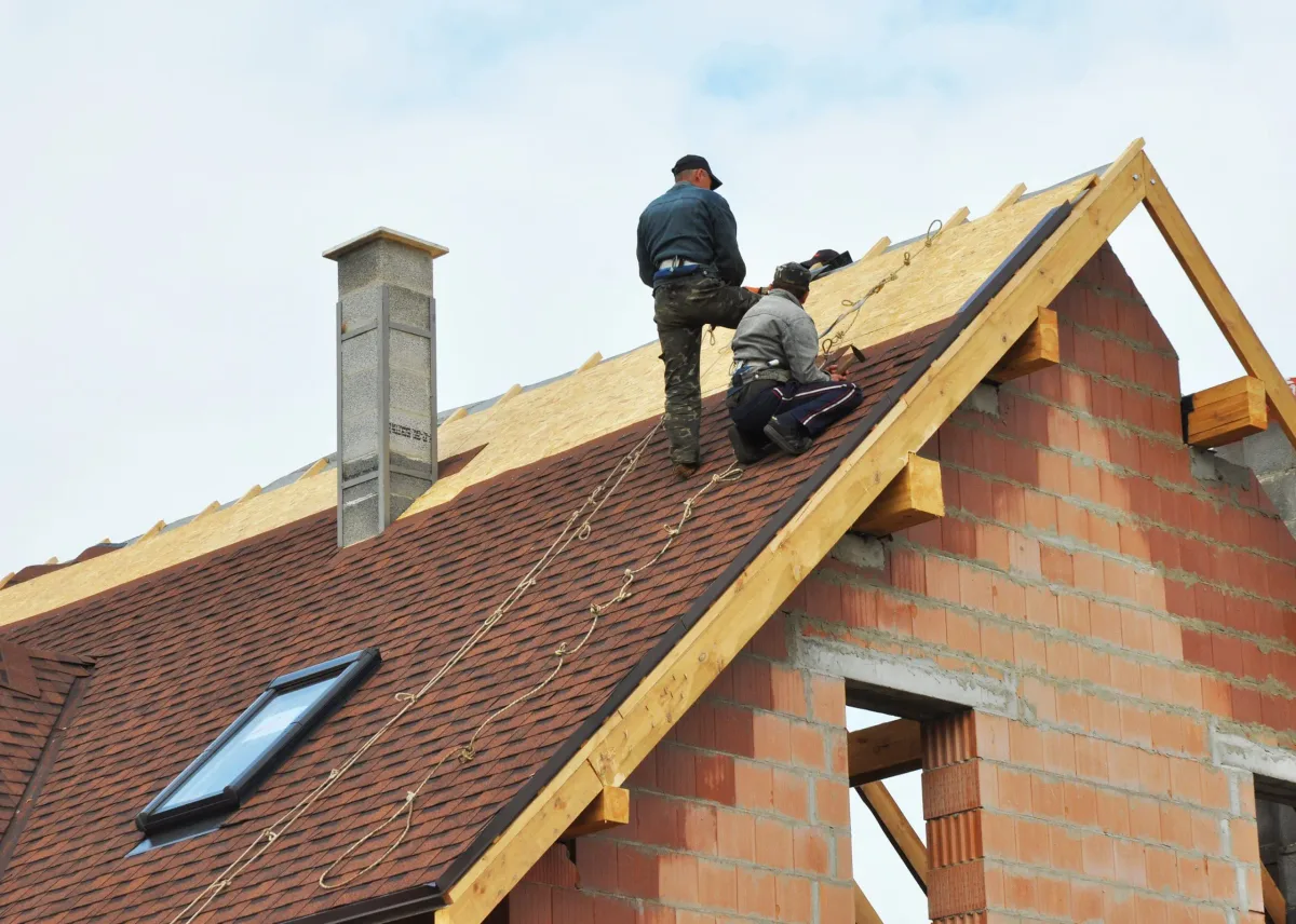 Two roofers with safety ropes shingling the roof of a brick house