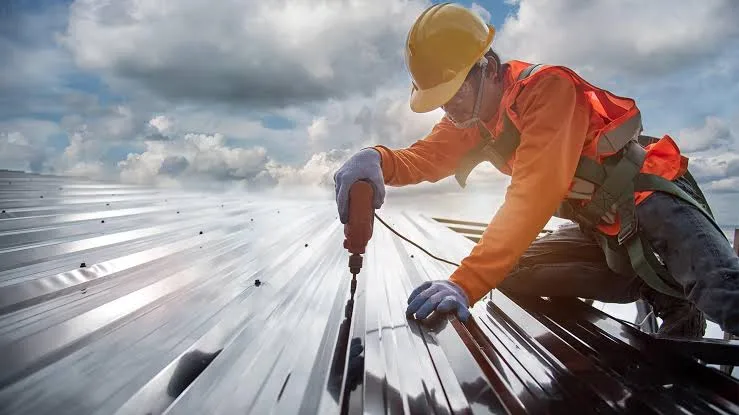 A roofer drilling a dark standing-seam metal roof under a dramatic sky