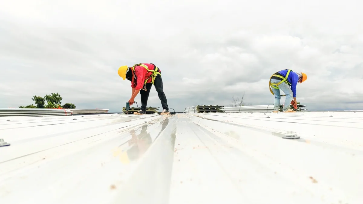 Two roofers fastening a white metal roof under an overcast sky