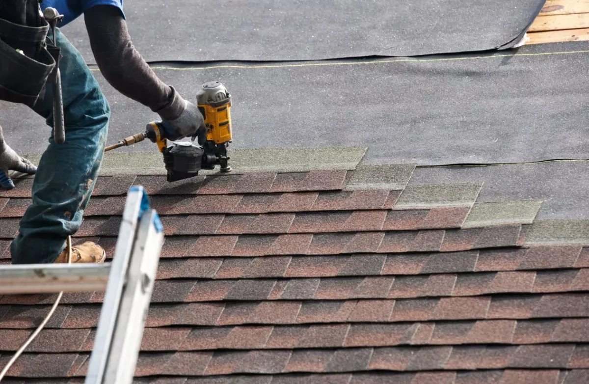 A roofer nailing red-brown shingles from a ladder