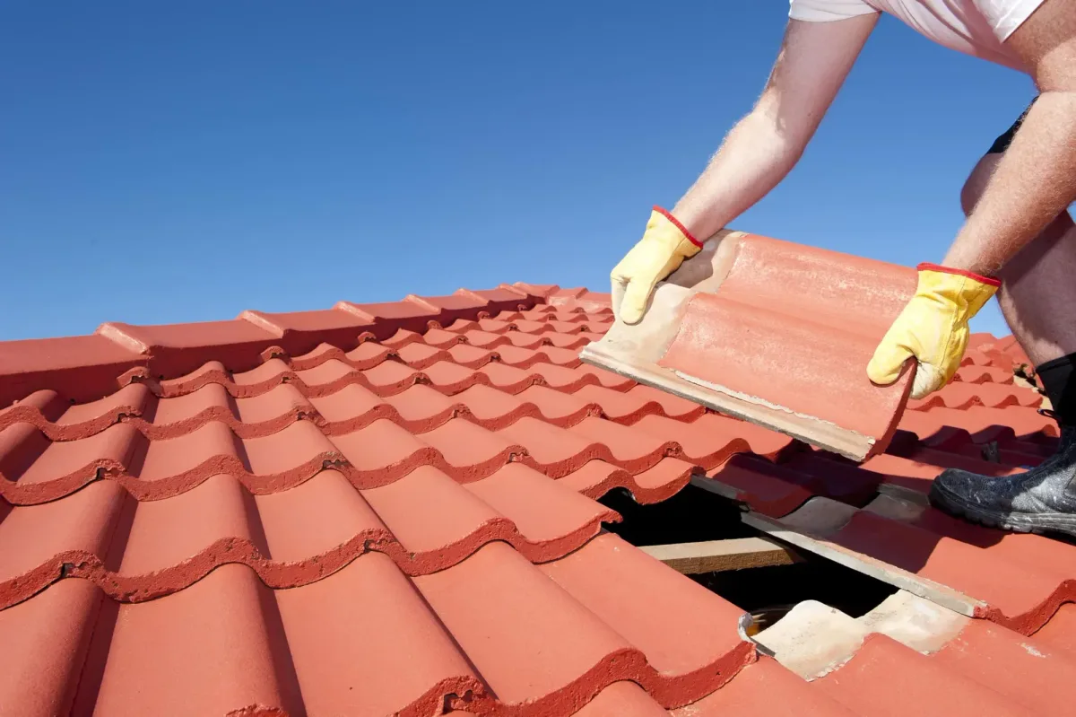 Gloved hands lifting a curved red clay roof tile against a blue sky