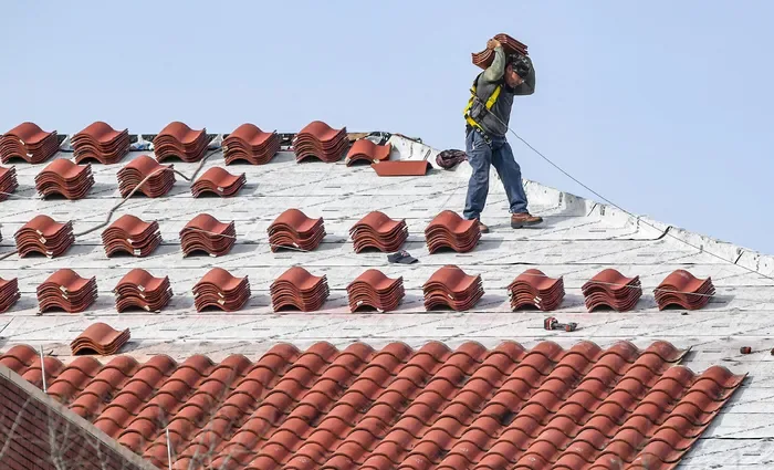 A roofer carrying a stack of red clay tiles across a roof deck