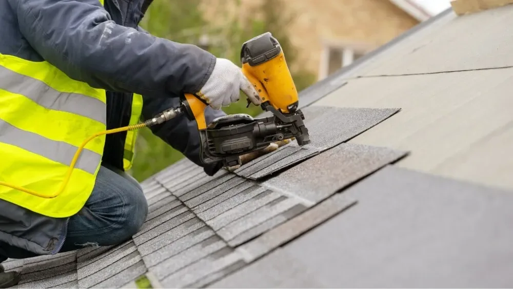A roofer in a high-visibility vest nailing shingles with a coil nailer